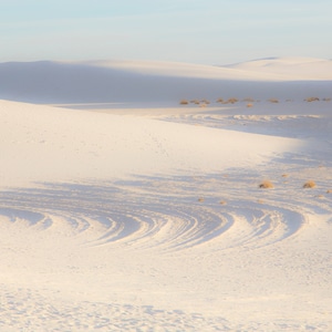 May include: A white sand dune with tire tracks and sparse vegetation. The sand is a bright white, and the sky is a pale blue.