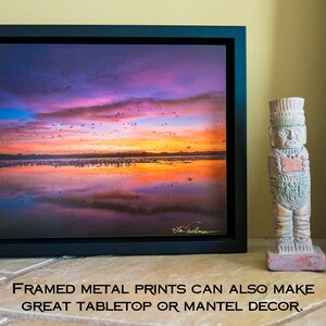 May include: A framed metal print of a colorful sunset over a lake with a flock of birds flying overhead. The print is on a table with two decorative statues.