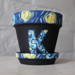 Puede incluir: Una maceta de terracota negra con un diseño de noche estrellada azul y amarilla. La maceta tiene un platillo a juego y la letra "K" pintada en la parte delantera.