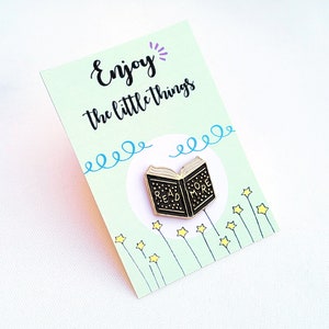 Peut inclure: Une épingle en émail noir et or en forme de livre ouvert avec les mots "READ MORE". L'épingle est sur une carte vert clair avec les mots "Enjoy the little things" et des illustrations d'étoiles. L'épingle mesure environ 2.5 cm de long.