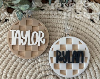 May include: Two round wooden ornaments with a checkered pattern. One ornament is brown with white text that says "Taylor". The other ornament is white with black text that says "Rylan".