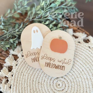 May include: Two wooden Halloween countdown signs. One features a white ghost, the other an orange pumpkin. Both signs have the text "sleeps until Halloween" in a script font. The signs are on a woven surface.