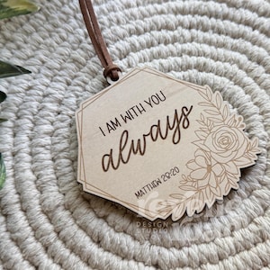 May include: A wooden plaque with the words "I AM WITH YOU always" and the Bible verse Matthew 28:20. The plaque has a floral design and a brown leather cord for hanging. The background is a woven, beige surface.