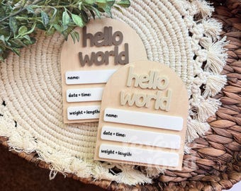 May include: Two wooden baby announcement plaques with the words "hello world" in raised letters. Each plaque has space to write the baby's name, date and time of birth, and weight and length. The plaques are light brown and beige.