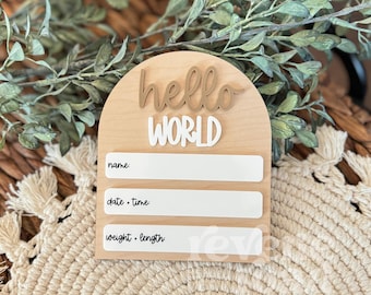 May include: Wooden arch-shaped sign with the words "hello world" in gold and white lettering. The sign has three blank lines for writing in the name, date and time, and weight and length of a newborn baby.