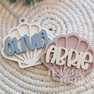 May include: Two seashell-shaped name tags. One is white with blue lettering spelling "Olivia", the other is pink with white lettering spelling "Abrie". Both are on a textured, off-white surface.