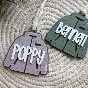 May include: Two jacket-shaped name tags. One is mauve with the name "POPPY" in white, the other is olive green with the name "Bennett" in white. Both tags have a cream-colored string attached for hanging.