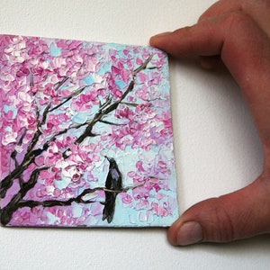 May include: A small painting of a black bird perched on a branch of a pink cherry blossom tree. The background is a light blue sky.