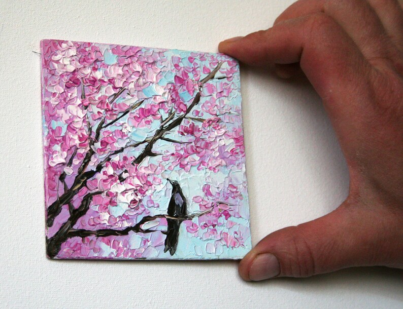May include: A small painting of a black bird perched on a branch of a pink cherry blossom tree. The background is a light blue sky.