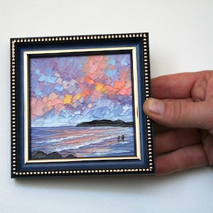 Puede incluir: Una pintura enmarcada que representa un paisaje marítimo con un cielo de puesta de sol colorido, agua azul y una pequeña isla en la distancia. La pintura está en un marco azul oscuro con detalles dorados.