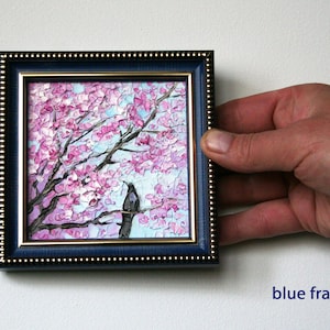 May include: A framed painting of a pink cherry blossom tree with a black bird perched on a branch. The frame is blue with a gold trim.
