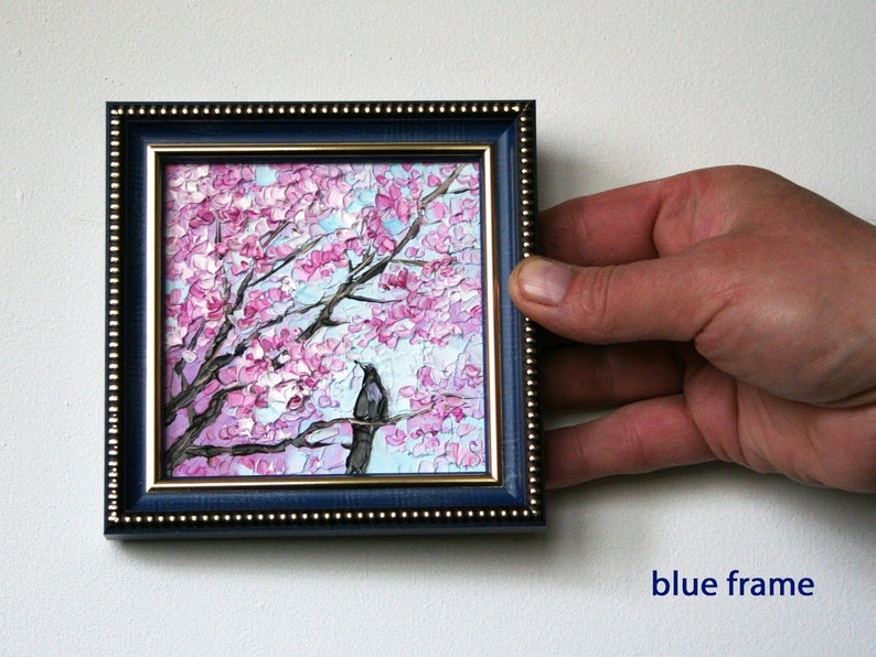 May include: A framed painting of a pink cherry blossom tree with a black bird perched on a branch. The frame is blue with a gold trim.