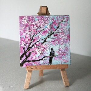 May include: A small painting on a wooden easel depicts a pink and white cherry blossom tree with a black bird perched on a branch. The painting is titled "TART".