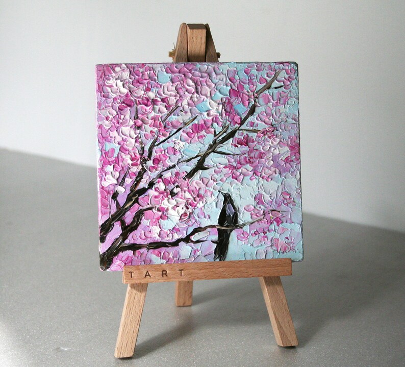 May include: A small painting on a wooden easel depicts a pink and white cherry blossom tree with a black bird perched on a branch. The painting is titled "TART".