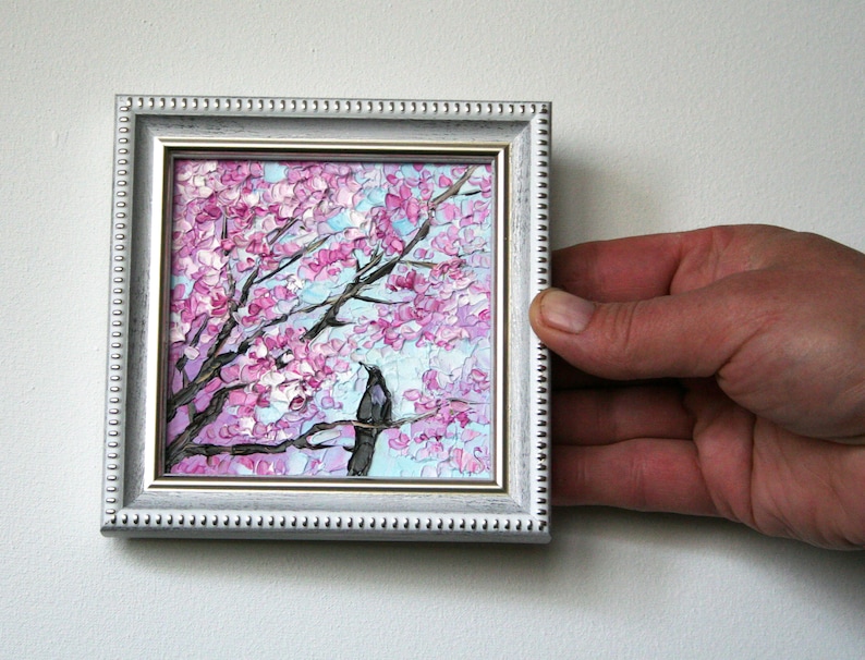 May include: A small framed painting of a pink cherry blossom tree with a black bird perched on a branch. The painting is in a white frame with a decorative border.
