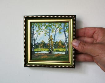 Op de afbeelding: Een kleine, ingelijste landschapsschilderij met berken met groene en gele bladeren in de buurt van een water. Het schilderij heeft een gouden en zwarte lijst. Het kunstwerk wordt door een hand tegen een witte achtergrond gehouden.