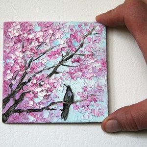 May include: A small painting of a black bird perched on a branch of a pink cherry blossom tree. The background is a light blue sky.