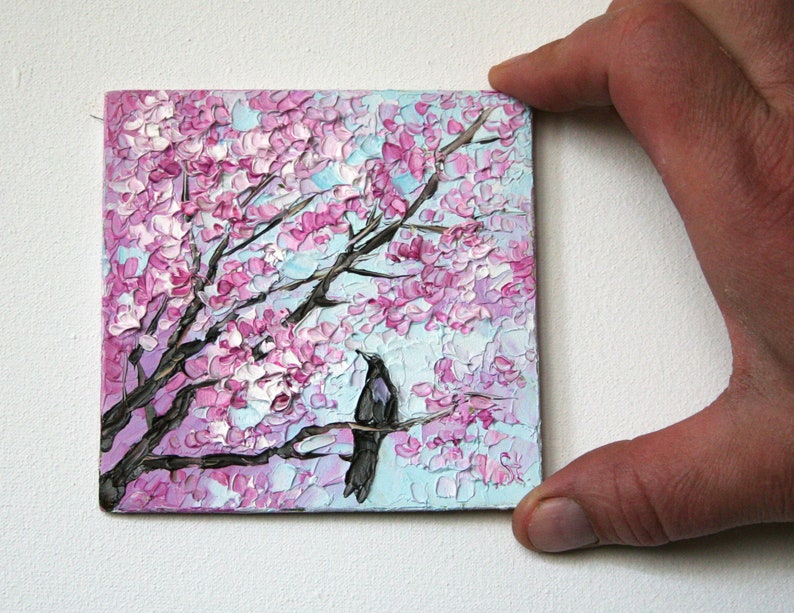 May include: A small painting of a black bird perched on a branch of a pink cherry blossom tree. The background is a light blue sky.