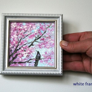 May include: A small painting in a white frame depicts a pink and purple cherry blossom tree with a black bird perched on a branch. The painting has a light blue background.