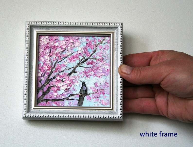May include: A small painting in a white frame depicts a pink and purple cherry blossom tree with a black bird perched on a branch. The painting has a light blue background.