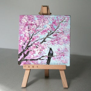May include: A small painting on a wooden easel depicts a pink and white cherry blossom tree with a black bird perched on a branch. The painting is signed "TART" in the bottom right corner.