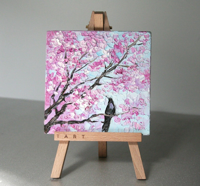 May include: A small painting on a wooden easel depicts a pink and white cherry blossom tree with a black bird perched on a branch. The painting is signed "TART" in the bottom right corner.
