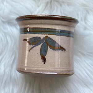 May include: A small, cream-colored ceramic pot with a brown rim and a blue and brown leaf design.
