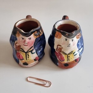 May include: Two small ceramic character jugs featuring hand-painted figures in blue jackets and yellow shirts. Each jug has a small handle and a pouring spout. A single copper-coloured metal paperclip is placed in front for scale reference.