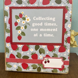 May include: A square scrapbook with a light blue cover featuring a repeating pattern of red strawberries, green leaves, and small white flowers. A central rectangular plaque displays the text, Collecting good times, one moment at a time.