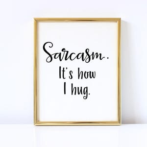 May include: A gold framed print with the text "Sarcasm. It's how I hug." in black cursive font on a white background.
