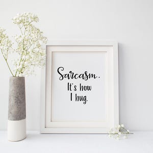 May include: White framed print with black text that reads "Sarcasm: It's how I hug." The print is sitting on a white surface with a vase of white flowers on the left.