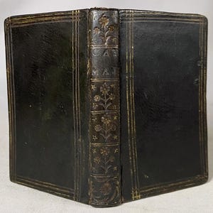 May include: A small, antique black leather-bound book featuring gold-tooled double-line borders on the front and back covers. The spine displays gold-embossed floral motifs and the letters CAT MAJ vertically aligned in a central panel.