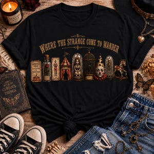May include: A black short-sleeved t-shirt featuring a graphic print with the text "WHERE THE STRANGE COME TO WANDER" above a row of circus-themed items including a ticket, a bottle, a tent, a doll in a glass dome, a birdcage, a clown, and a skeleton.