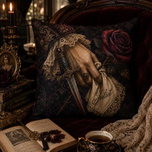 Puede incluir: Un cojín decorativo cuadrado con un patrón de damasco en color morado oscuro y negro. El gráfico central muestra una mano con puño de encaje sosteniendo una daga y una rosa roja, junto a un pañuelo de encaje bordado con el nombre Juliet.