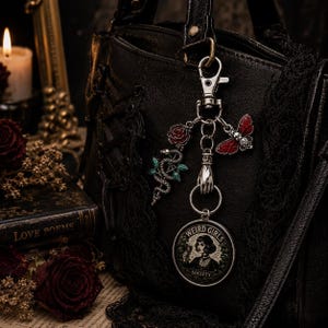 Op de afbeelding: Een zilverkleurige metalen sleutelhanger met een ronde bedel met de tekst "WEIRD GIRLS SOCIETY" en een afbeelding van een persoon die een glas vasthoudt. De sleutelhanger bevat bedels van een rode roos, een groen-zilveren slang, een rood insect en een hand.