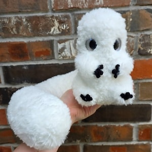 May include: A plush toy made of white fuzzy material with a long tail, a rounded head, and large dark eyes. The creature features small black stitched details on its front and back paws, giving it a distinct appearance with a soft texture.