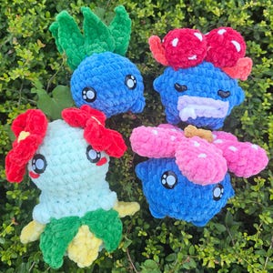 May include: Four handmade crochet plush toys in various colours. The toys include a blue creature with green leaves, a blue creature with red mushroom-like features, a light green creature with red flower-like features, and a blue creature with pink flower-like features.