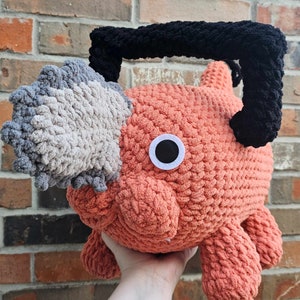 Könnte beinhalten: Ein gehäkeltes Plüschtier in Form einer Kreatur mit einem großen, grau-weißen Ohr, einem leuchtend orangefarbenen Körper und einem schwarzen gehäkelten Stirnband. Das Spielzeug hat ein einzelnes weißes Auge mit einer schwarzen Pupille.