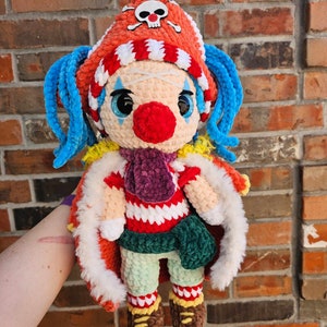 May include: A crocheted plush toy with a red and white striped hat, blue hair, and a red nose. The toy is wearing a green and red striped shirt and a purple scarf.