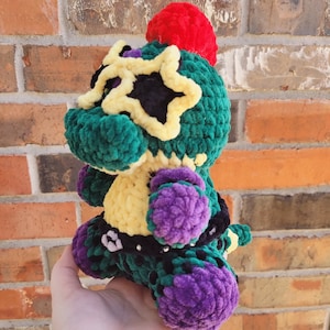 Könnte beinhalten: Ein grünes, gelbes und violettes gehäkeltes Plüschtier mit einem roten Bommel auf dem Kopf. Das Spielzeug trägt einen schwarzen Gürtel mit einer silbernen Schnalle und hat ein sternförmiges Design auf dem Gesicht.