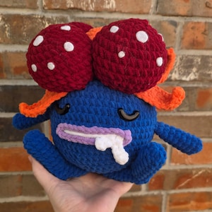 Puede incluir: Una criatura azul hecha a mano con dos cabezas rojas en forma de hongo, cada una con puntos blancos. Tiene detalles en naranja, ojos negros curvos, una boca morada y baba blanca. El peluche se sostiene en una mano.