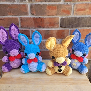 Könnte beinhalten: Vier gehäkelte Plüschtiere in Form von Hasen. Die Hasen sind lila, blau, braun und blau. Jeder Hase hat eine Schleife und gestickte Augen.