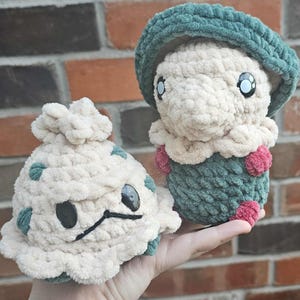 May include: Two plush crochet mushroom figures. One is a cream color with green spots and a black stitched face. The other is held in a hand and has a green hat, cream body, and pink accents. Both have large black button eyes.
