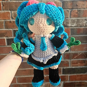 May include: A handmade crochet doll with blue hair, eyes, and accents. The doll wears a gray and black outfit with blue trim and green accents. The doll is held in front of a brick wall.