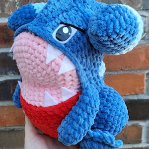 May include: A blue and pink crocheted plush toy shaped like a shark with a toothy grin. The toy has a white eye and a red belly.