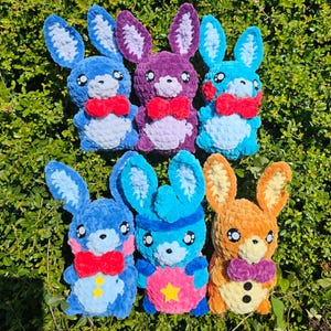 May include: Six handmade plush bunnies in various colors, including blue, purple, and orange. Each bunny has large ears, a bow tie, and embroidered facial features. Some bunnies have star or circle accents. The bunnies are arranged on a bed of green foliage.