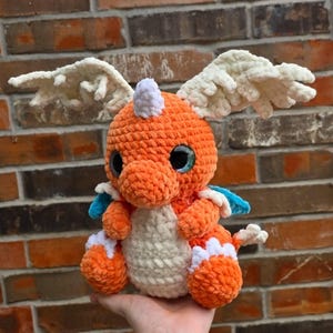 May include: A handmade orange crochet dragon plush toy with white wings, a white belly, and blue accents. The dragon has large, black eyes with a hint of blue and a white horn. The plush toy is held in a hand.