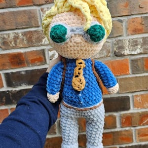 May include: A crocheted plush toy of a person with yellow hair, green goggles, a blue shirt, a brown tie, and gray pants.