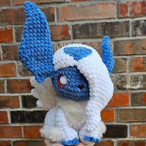 May include: A handmade plush toy of a character with blue and white fur, large blue ears, and a red and black eye. The plush has a soft, textured appearance and is set against a brick background.