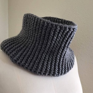 Peut inclure: Un chauffe-cou gris en tricot côtelé. Le chauffe-cou est fait d'un fil épais et doux et présente un design large et évasé. Le col est présenté sur un mannequin blanc, mettant en valeur sa forme et sa texture. Cet accessoire est conçu pour garder le cou au chaud.